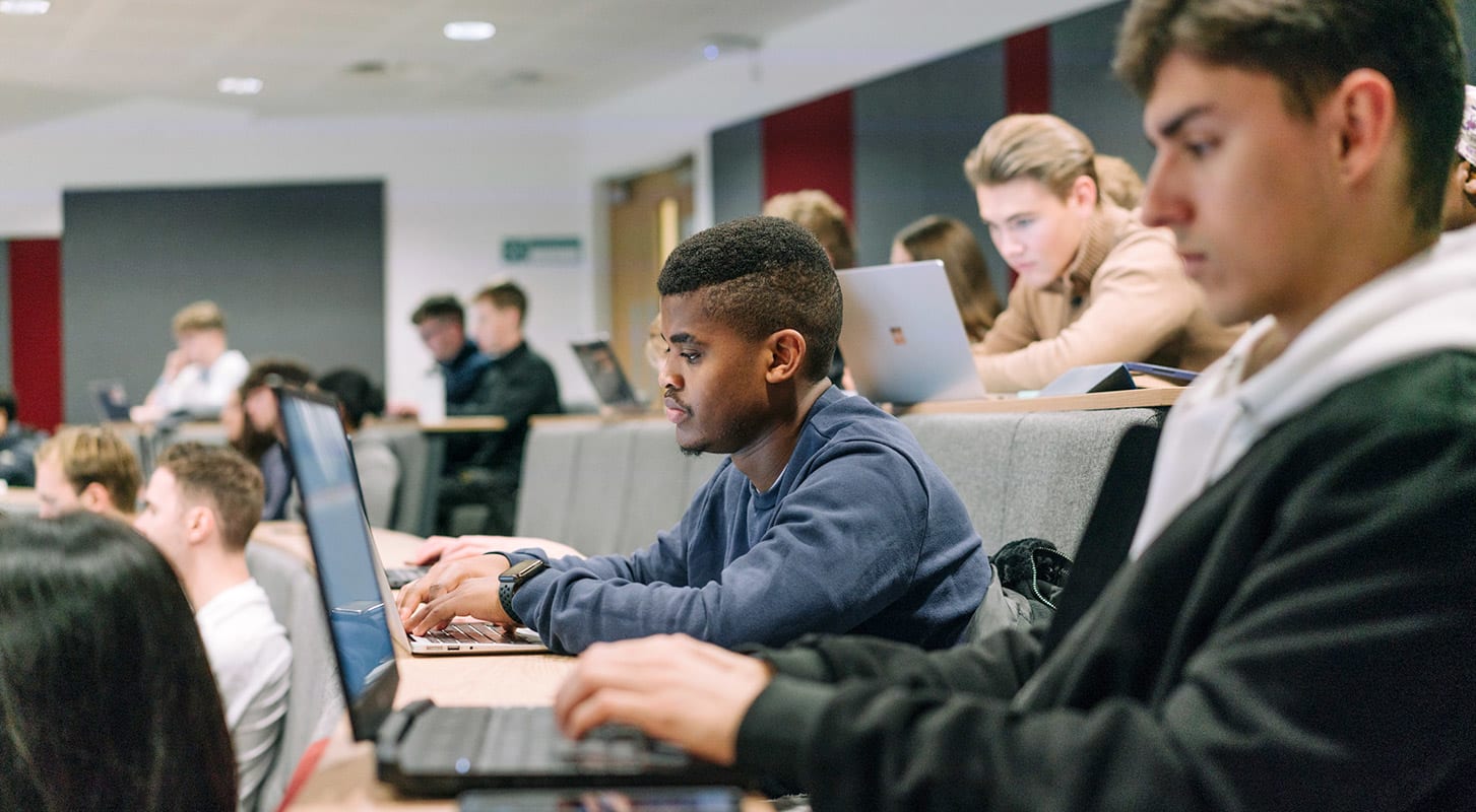 A diverse group of students in a lecture hall focused on their laptops during a lecture.